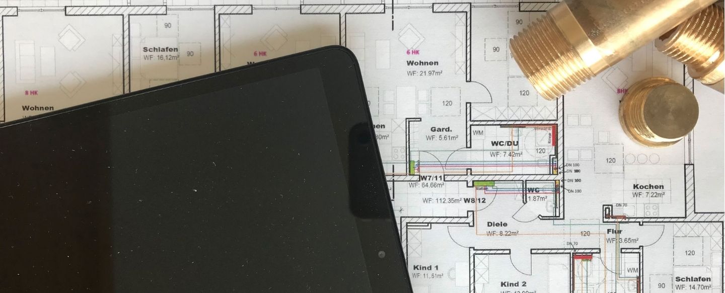 Ein technischer Bauplan einer Wohnung liegt auf einem Tisch und zeigt detaillierte Raumaufteilungen. Ein schwarzes Tablet verdeckt teilweise den linken Bereich des Plans, während auf der rechten Seite mehrere Messingrohrstücke liegen. Die Kombination von Tablet, Bauplan und Rohrverbindungsstücken suggeriert eine Planung oder Überprüfung von Installationsarbeiten, möglicherweise im Bereich Heizung, Lüftung oder Sanitär.