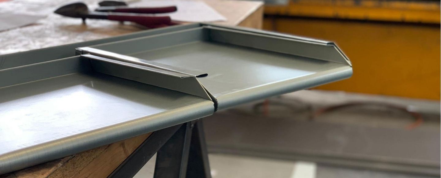 Ein naher Blick auf ein fertiggestelltes Metallblech, das auf einem Arbeitstisch liegt. Das Blech ist präzise gefaltet und zeigt Kanten, die möglicherweise Teil einer Dach- oder Fassadenverkleidung sind. Im Hintergrund liegen Werkzeuge, darunter eine Zange und andere Arbeitsutensilien, die andeuten, dass das Blech gerade in einer Werkstatt bearbeitet wurde. Die Szene vermittelt den Eindruck handwerklicher Präzision und technischer Arbeit.