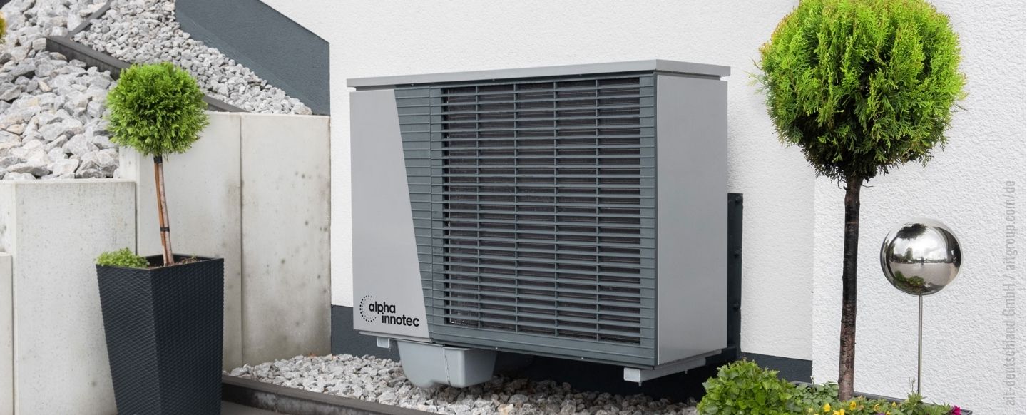 Eine Außeneinheit einer Wärmepumpe der Marke 'alpha innotec' ist an der Außenwand eines Gebäudes installiert. Die graue, rechteckige Einheit steht auf einem Kiesbett und ist von moderner Gartengestaltung umgeben, darunter zwei gepflegte Zierpflanzen in schwarzen Töpfen und eine metallische Kugeldekoration. Im Hintergrund sind Betonwände und eine mit Steinen bedeckte Böschung zu sehen.