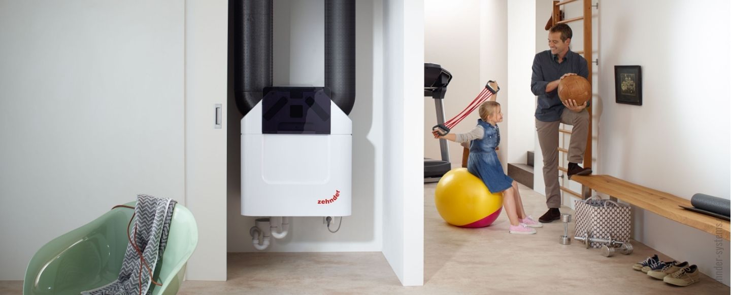 In einem modern eingerichteten Raum ist ein Lüftungssystem der Marke 'Zehnder' an der Wand installiert. Das weiße Gerät mit schwarzen Rohrleitungen ist zentral in einer Nische platziert. Im Hintergrund spielt ein kleines Mädchen auf einem gelben Gymnastikball, während ein Mann, vermutlich ihr Vater, mit einem Basketball in der Hand neben ihr steht. Der Raum scheint ein Fitness- oder Hobbyraum zu sein, mit Sportgeräten wie einem Laufband, einer Sprossenwand und Hanteln. Auf einer Bank an der Wand liegen Sportaccessoires, während im Vordergrund ein grüner Stuhl mit einer Decke darauf zu sehen ist.