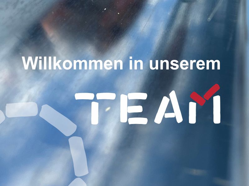 Ein Bild mit der Aufschrift „Willkommen in unserem TEAM“ vor einem unscharfen, metallisch wirkenden Hintergrund. Das Wort „TEAM“ ist stilisiert, wobei das „M“ durch ein rotes Häkchen akzentuiert ist.