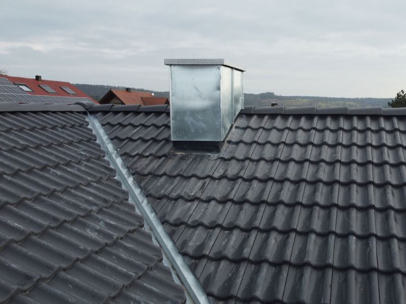 Ein graues Ziegeldach mit einem rechteckigen, silbernen Schornstein in der Mitte. Das Dach besteht aus wellenförmigen Dachziegeln, und im Hintergrund sind mehrere Häuser sowie der Himmel zu sehen.