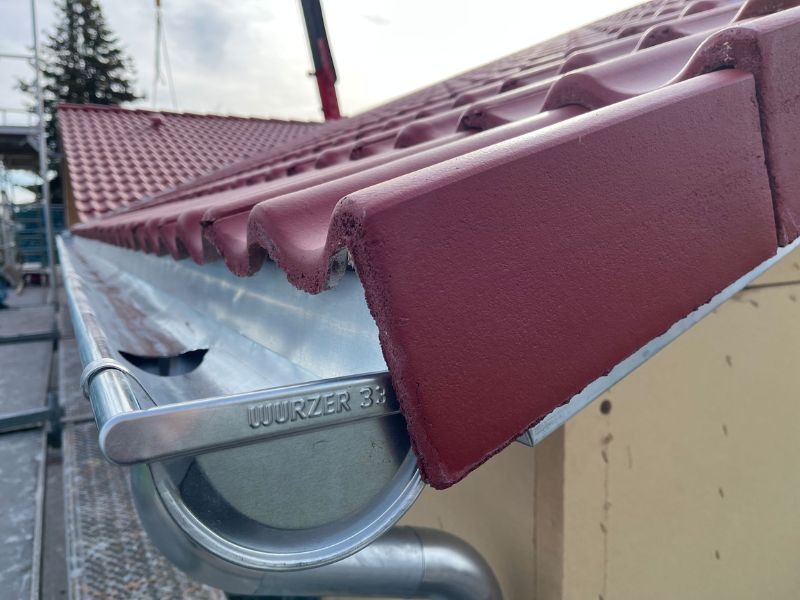 Nahaufnahme einer Dachrinne, die unter einem Dach mit roten Ziegeln montiert ist. Die Ziegel haben eine gewellte Struktur, und die silberne Metallrinne trägt die Prägung „Würzer 333“.