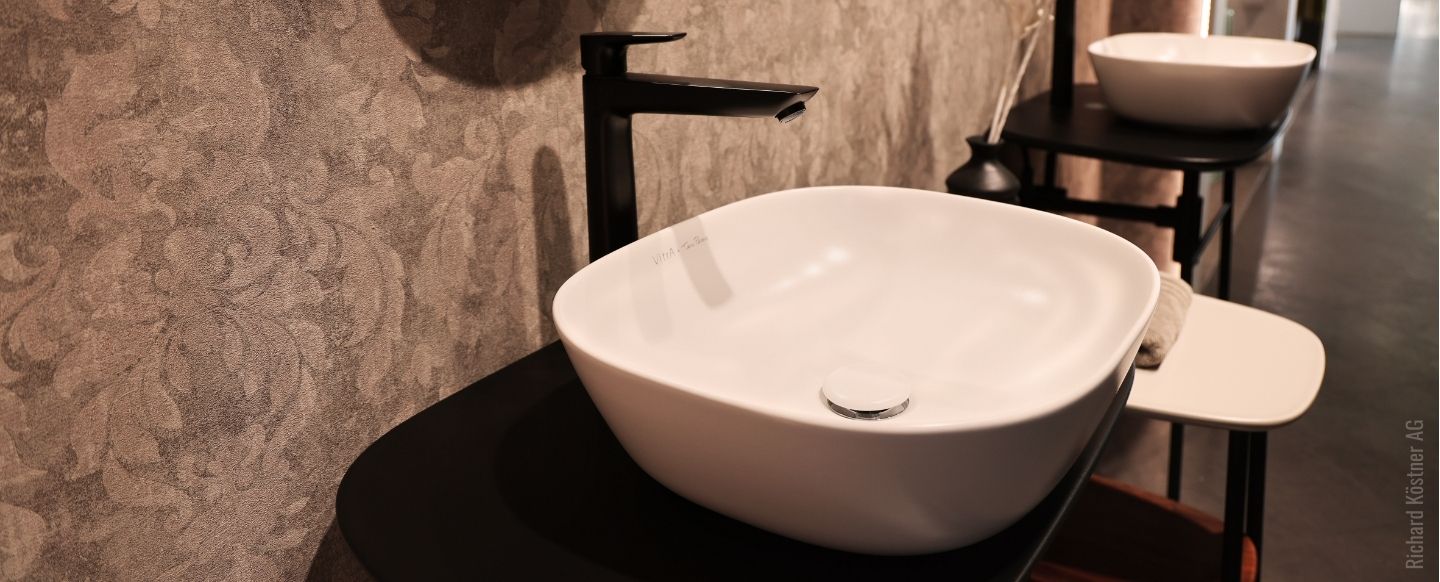 Ein stilvoll eingerichtetes Badezimmer mit einem modernen, weißen Aufsatzwaschbecken, das auf einer schwarzen, minimalistischen Konsole montiert ist. Das Waschbecken hat eine ovale Form und wird von einem eleganten, mattschwarzen Wasserhahn ergänzt, der an der Rückseite des Beckens angebracht ist. Im Hintergrund ist ein weiteres identisches Waschbecken auf einer ähnlichen Konsole zu sehen. Die Wand im Hintergrund ist mit einer strukturierten, floral gemusterten Tapete in einem dezenten Grauton verziert, die dem Raum eine elegante und warme Atmosphäre verleiht. Foto: Richard Köstner AG