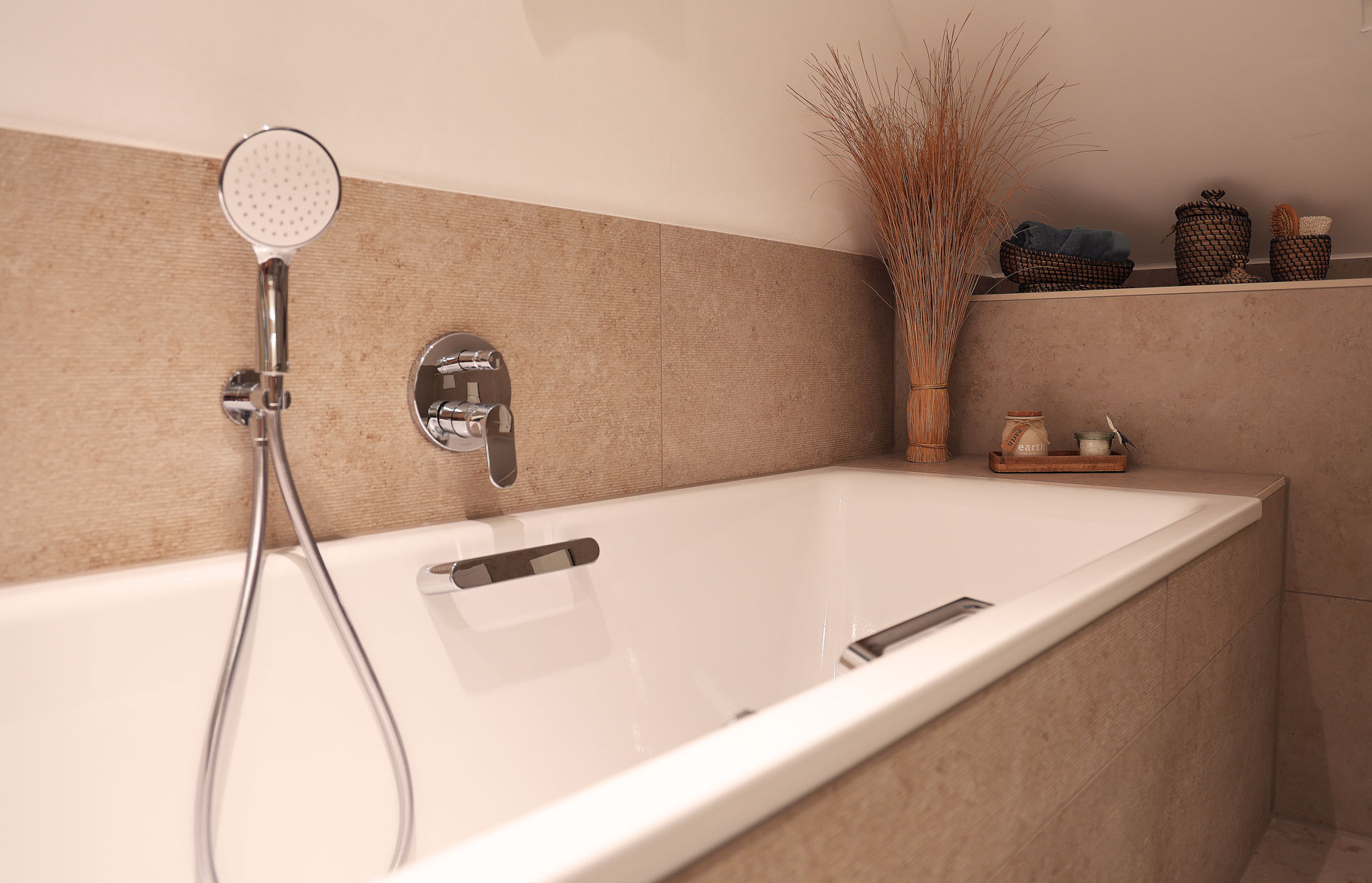 Ein modernes Badezimmer mit einer großen, rechteckigen Badewanne. An der Wand befindet sich eine verchromte Handbrause mit einem Mischhebel, während im Hintergrund eine dekorative Vase mit getrockneten Gräsern und einige kleine Accessoires auf einem Regal zu sehen sind. Foto: Richard Köstner AG 