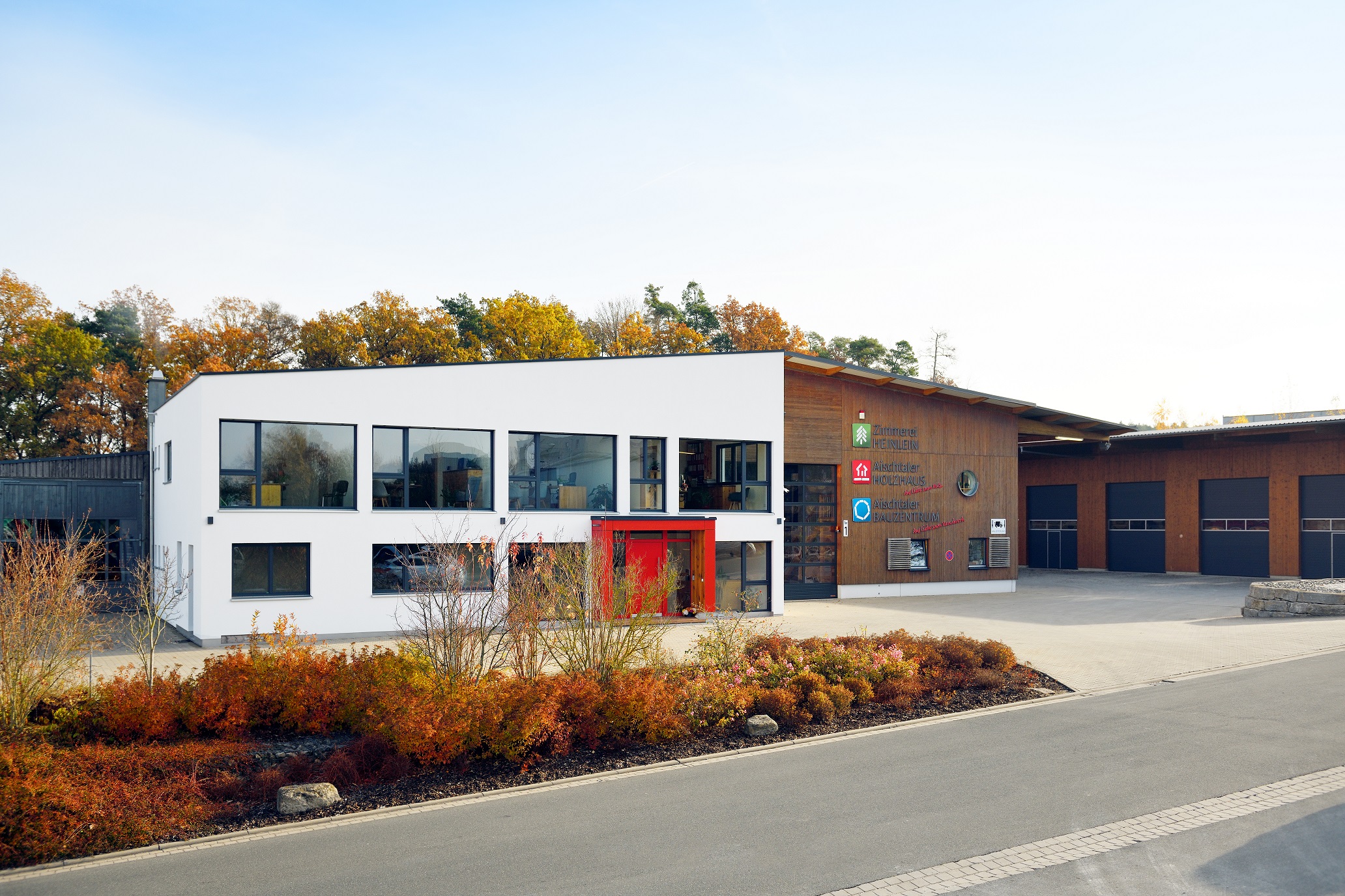 Ein zweistöckiges Gebäude mit einer modernen Architektur, das aus einem weißen und einem holzverkleideten Teil besteht. Die linke Hälfte des Gebäudes ist in schlichtem Weiß gehalten und hat große Fensterfronten, während die rechte Hälfte eine warme Holzverkleidung aufweist, die mit verschiedenen Firmenlogos verziert ist. Vor dem Gebäude erstreckt sich ein Beet mit herbstlich gefärbten Sträuchern. Der Hintergrund wird von Bäumen mit goldenen und orangenen Blättern dominiert, die unter einem klaren Himmel leuchten.