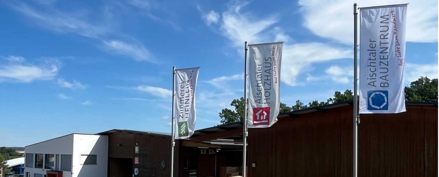 Mehrere hohe Fahnenmasten mit weißen Flaggen, die im Wind wehen, stehen vor einem modernen Gewerbegebäude. Auf den Flaggen sind die Logos und Schriftzüge der Unternehmen 'Zimmerei Heinlein', 'Aischtaler Holzhaus' und 'Aischtaler Bauzentrum' zu sehen. Im Hintergrund ist ein Teil des Gebäudes mit einer weißen und einer holzverkleideten Fassade sichtbar, während der Himmel darüber klar und blau ist, mit einigen weißen Wolken.